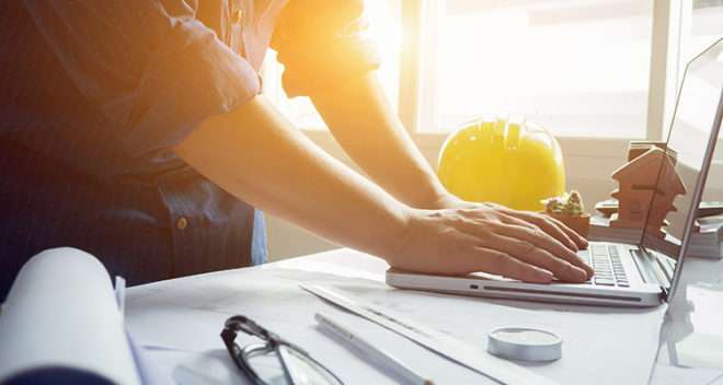 Person working on a laptop next to some notes and a construction helmet.